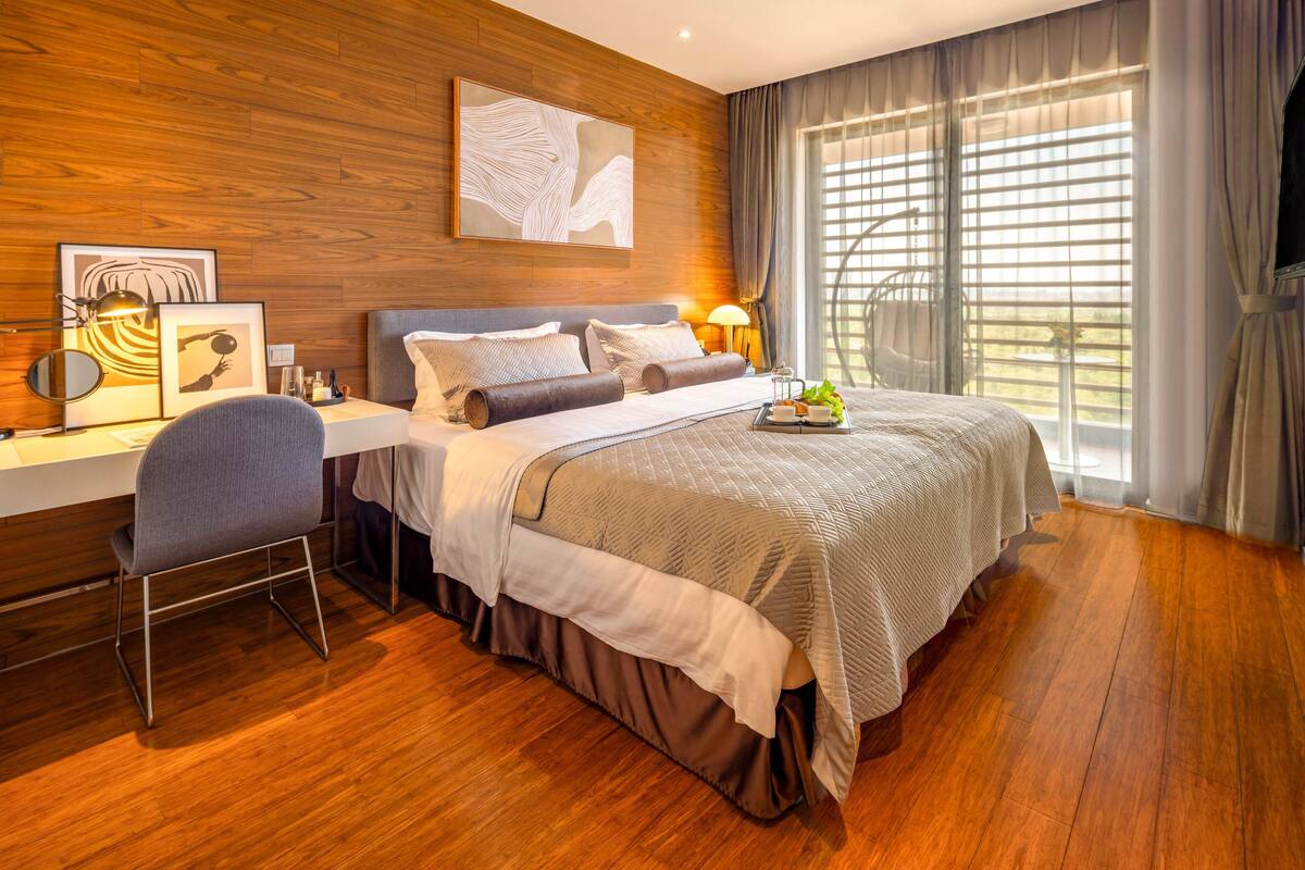A modern hotel room with a wooden floor, a large bed with a quilted duvet, a desk with a chair, a bedside table with a lamp, and a large window with curtains.