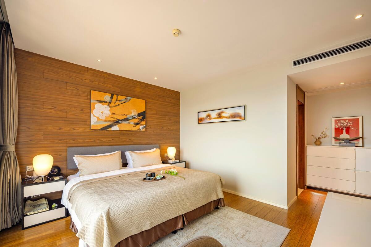 A modern bedroom with a wooden headboard, a large bed with a beige quilt, bedside tables with lamps, and artwork on the walls.