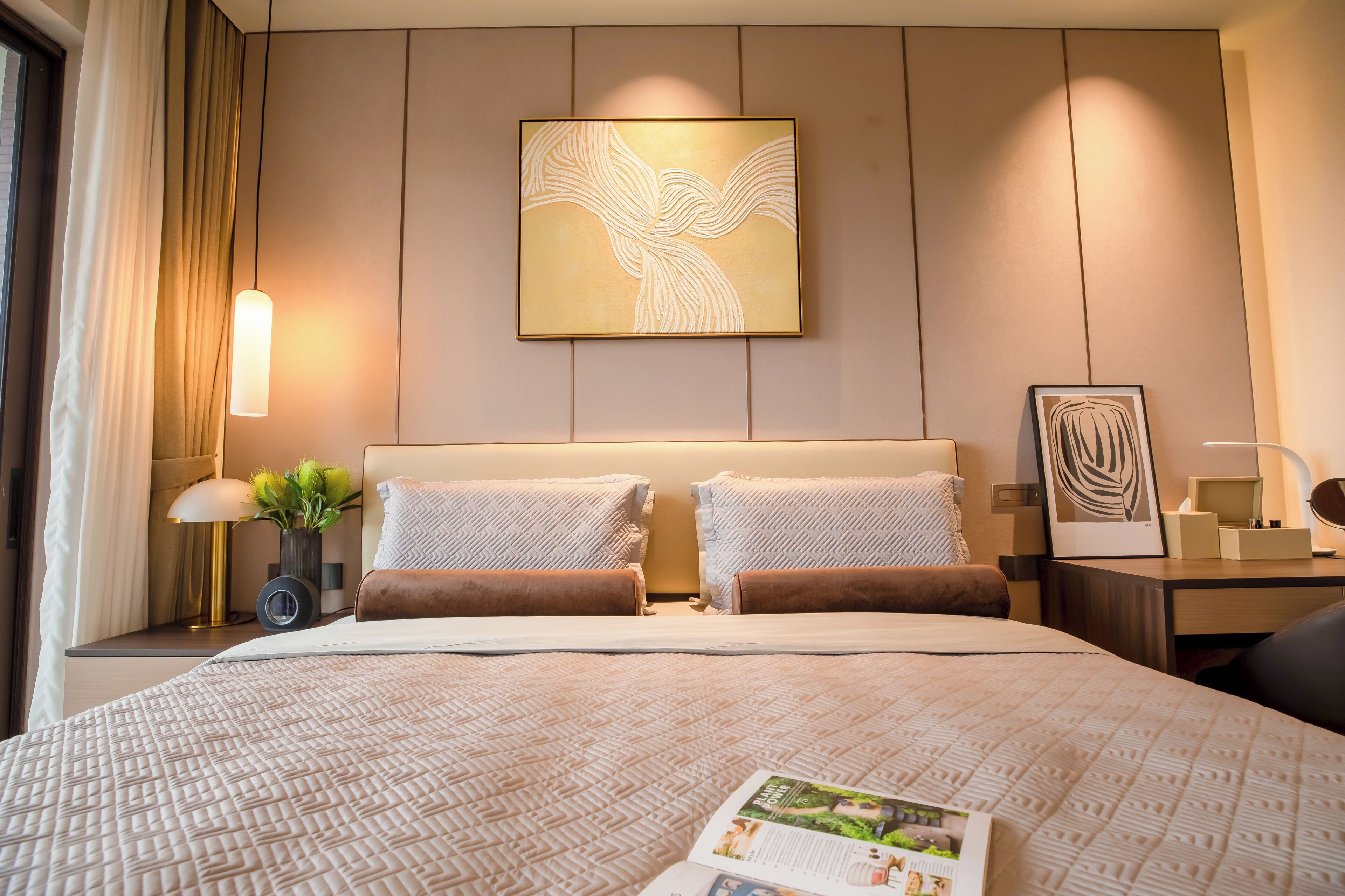Hotelkamer met een groot bed, twee kussens, een nachtkastje met een lamp, een kunstwerk in een lijst aan de muur, en een tijdschrift op het bed.