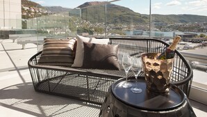 A balcony with a wicker sofa, a metal table with a bottle and glasses, and a view of a town and mountains.