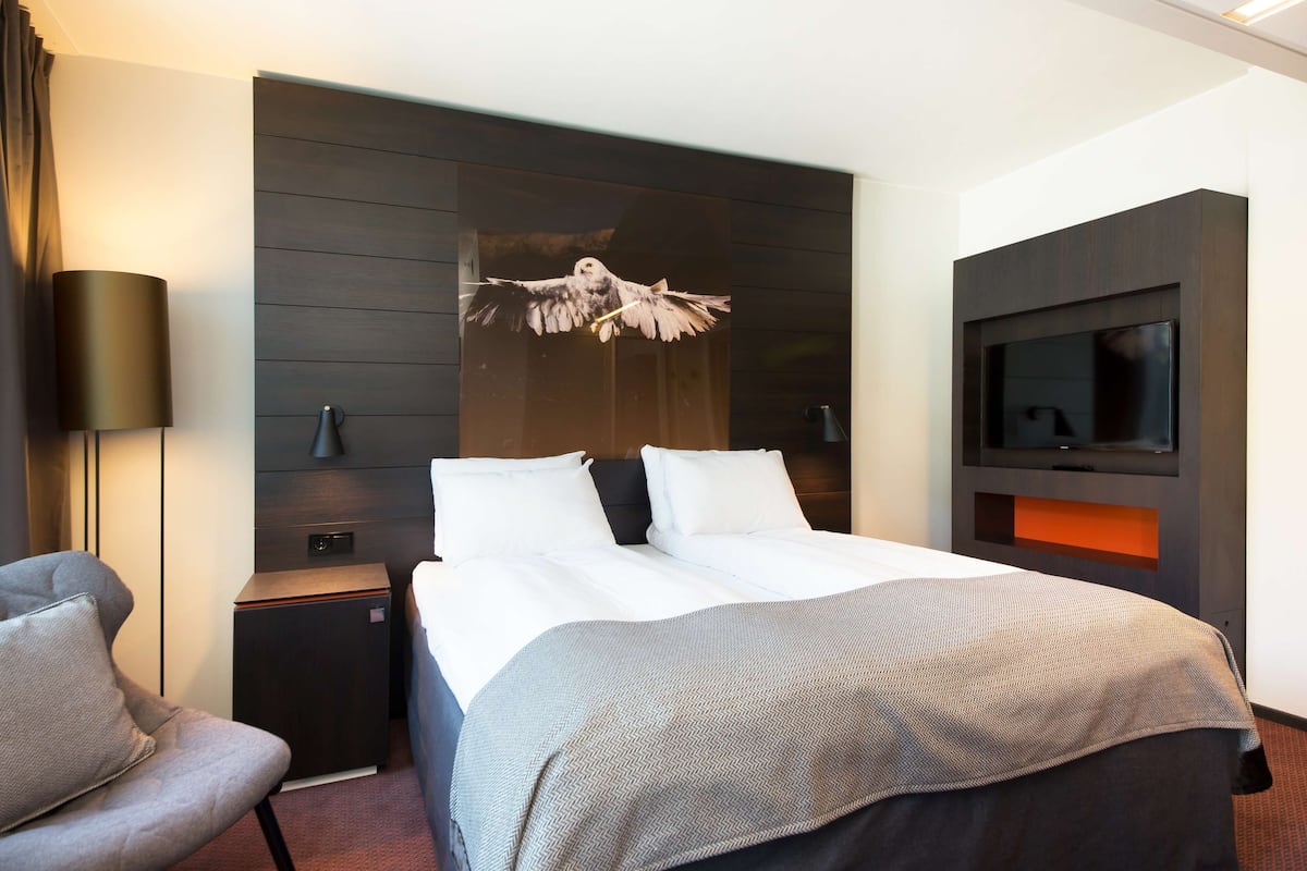 A modern hotel room with a large bed, a flat-screen TV, a desk with a lamp, and a chair.