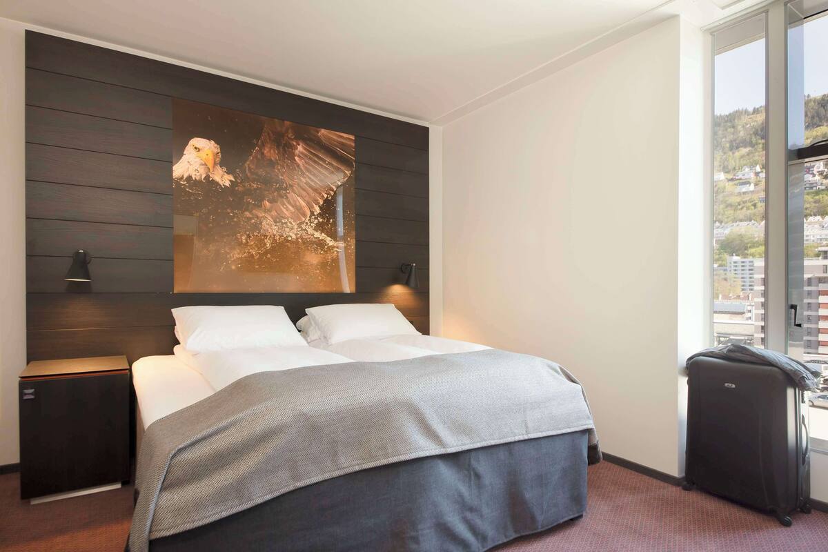 A hotel room with a bed, a nightstand, a lamp, a suitcase, and a large window with a city view.