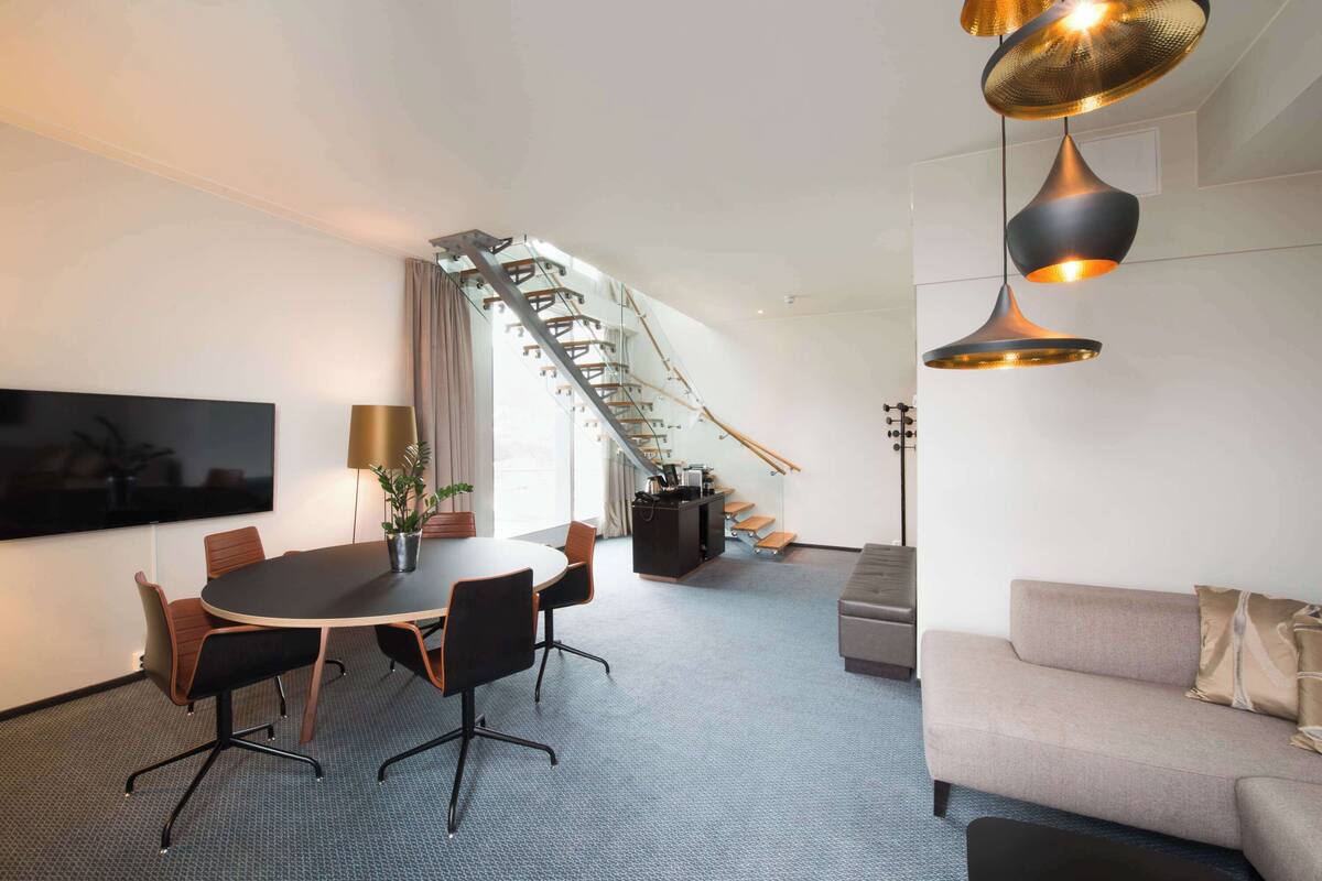 A modern hotel room with a conference table, sofa, staircase, and a TV on the wall.