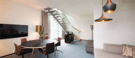 A modern hotel room with a conference table, sofa, staircase, and a TV on the wall.