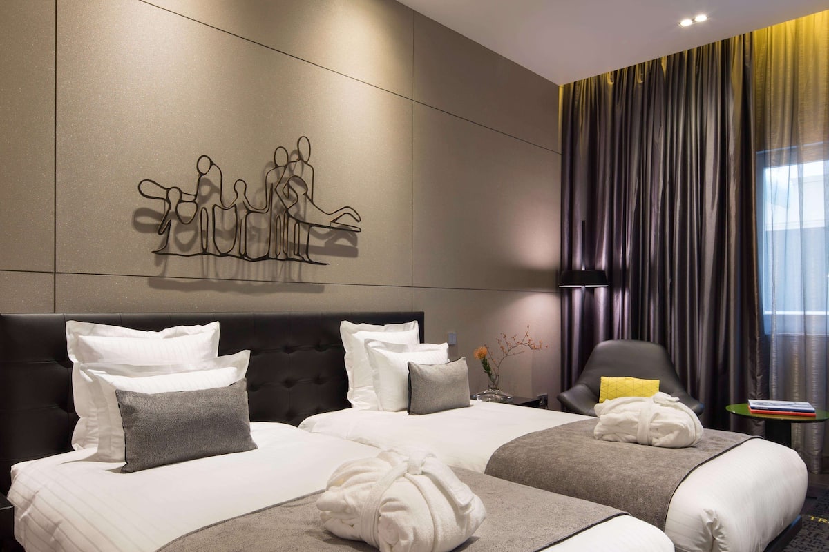 Ein Hotelzimmer mit zwei Betten, einem modernen Kopfteil, einem Wandbild, einem Fenster mit Vorhängen und einem kleinen Tisch mit einem Buch.