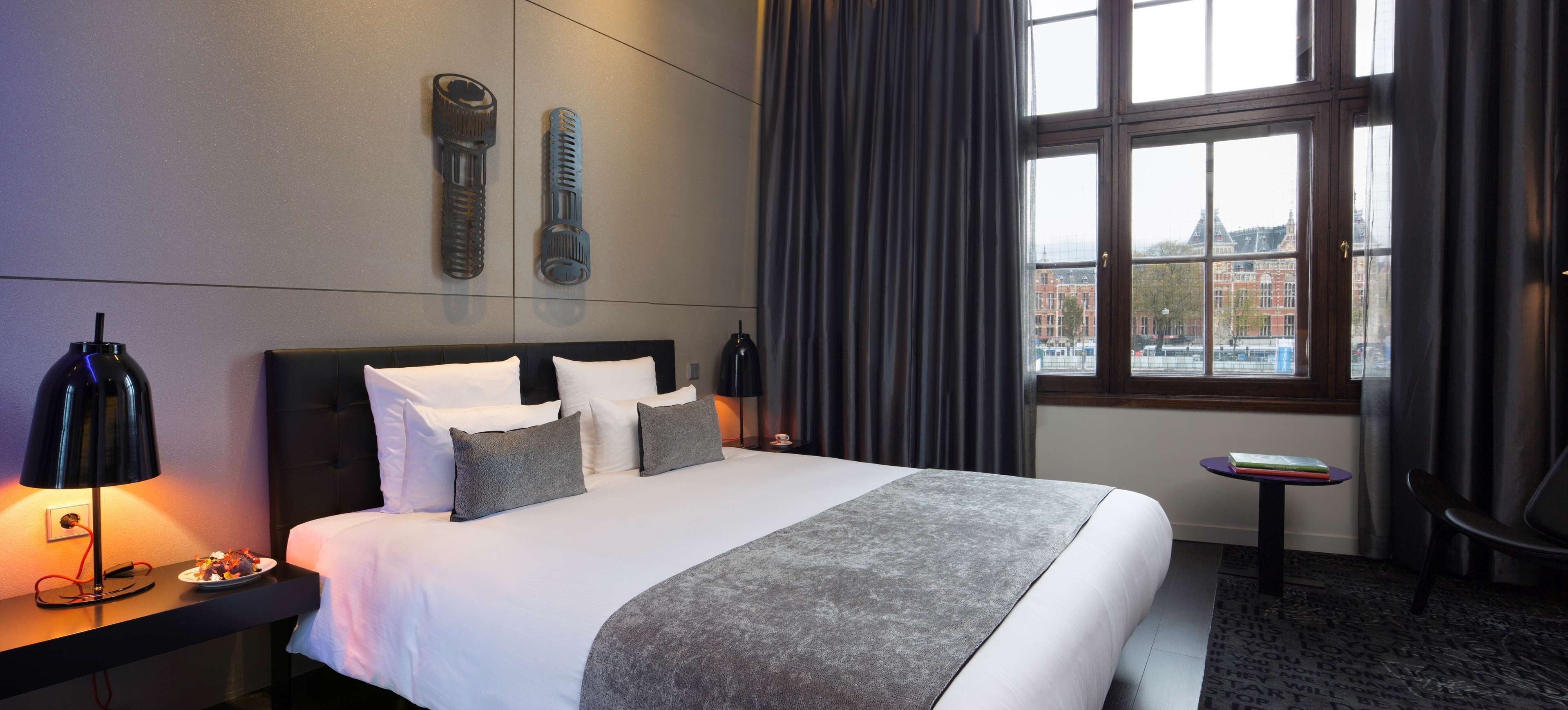 A hotel room with a large bed, two wall-mounted lamps, a small table with a book, and a view of buildings through the window.