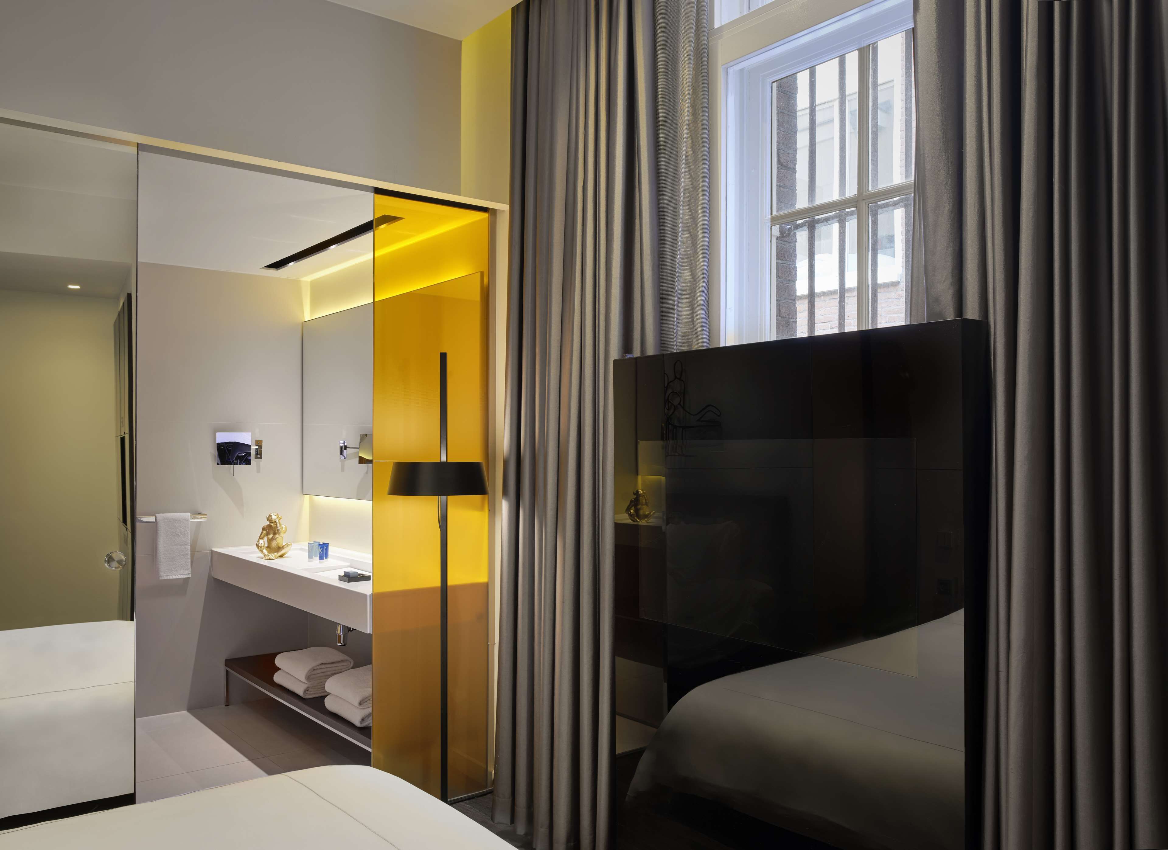 A hotel room with a bed, a bedside table, a mirror, a lamp, and a window with curtains.