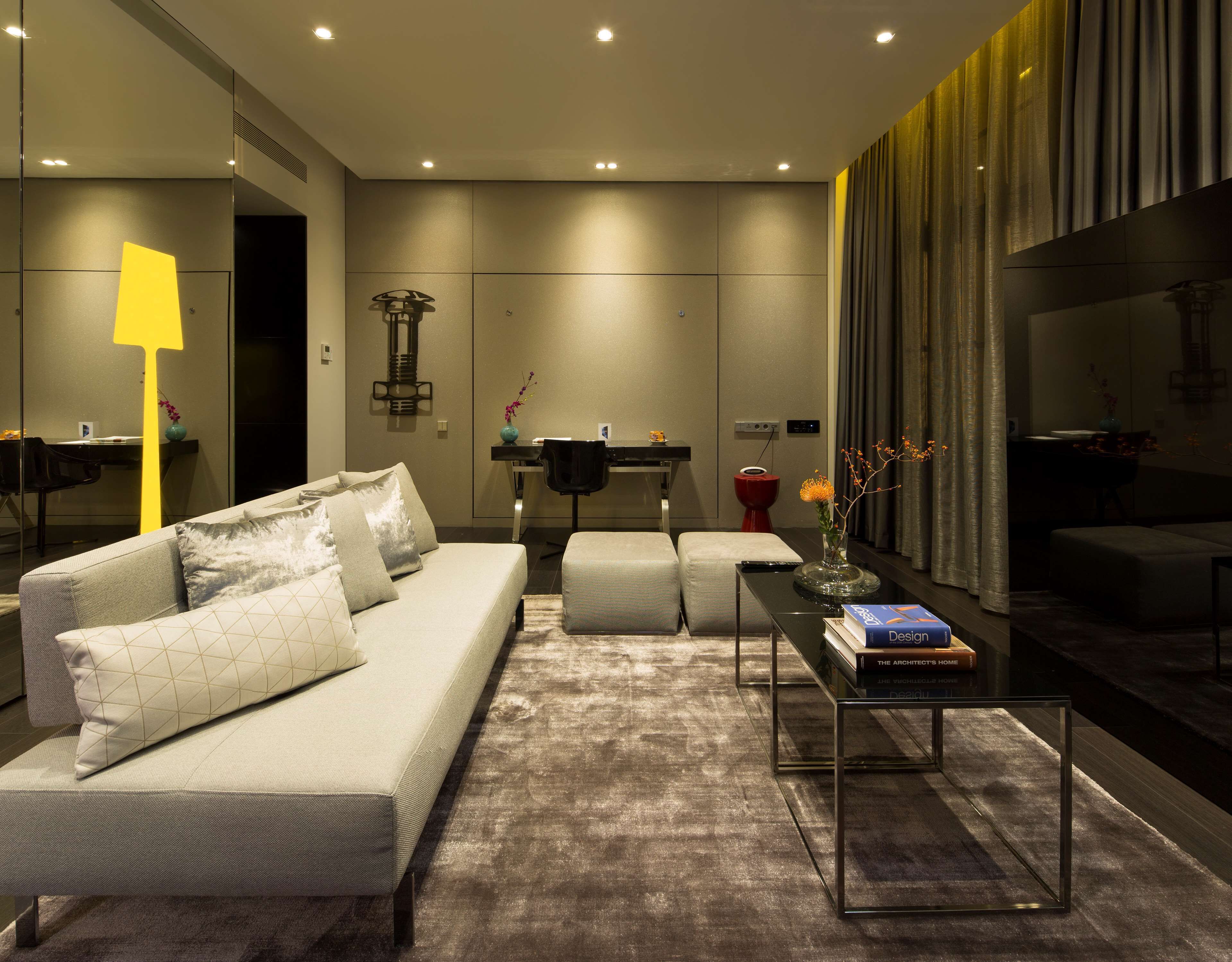 Un salon moderne avec un canapé, un pouf et une table basse. On y trouve également un coin repas avec une table et des chaises, ainsi qu’une grande fenêtre agrémentée de rideaux.
