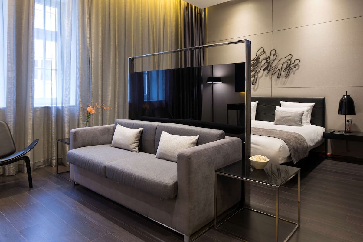 Ein modernes Hotelzimmer mit grauem Sofa, einem großen Flachbildfernseher, einem Bett mit weißer Bettwäsche und einem an der Wand hängenden Kunstwerk.
