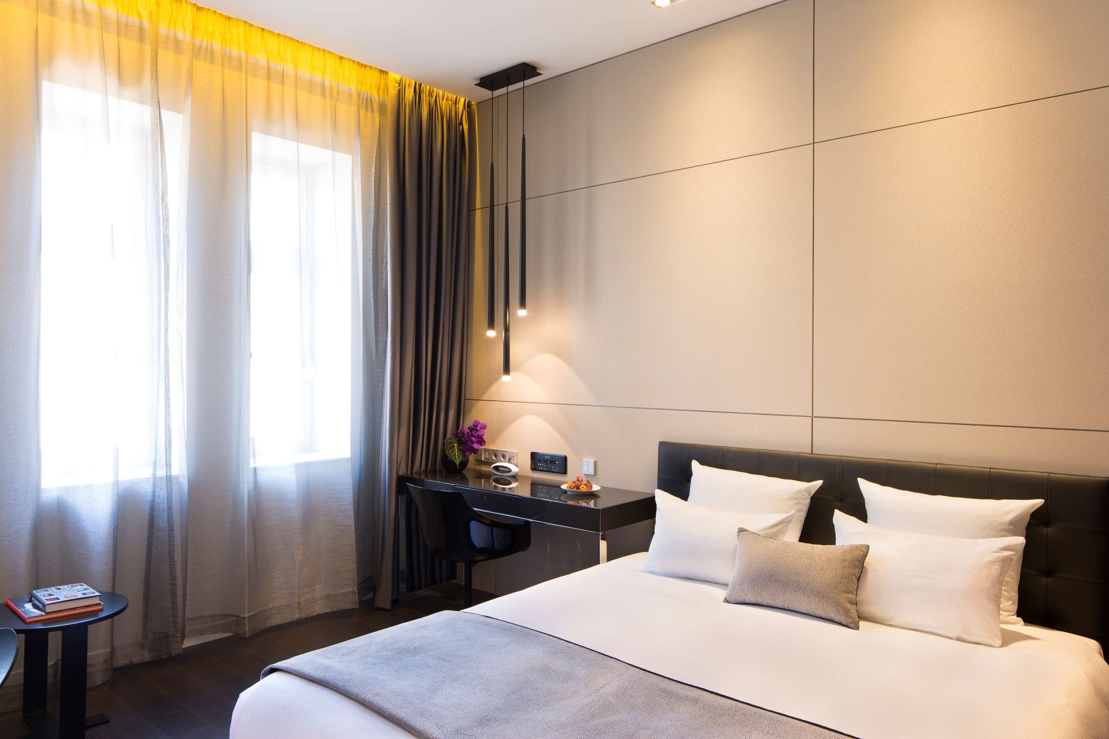 A modern hotel room with a large bed, a desk with a chair, and a window with sheer curtains.
