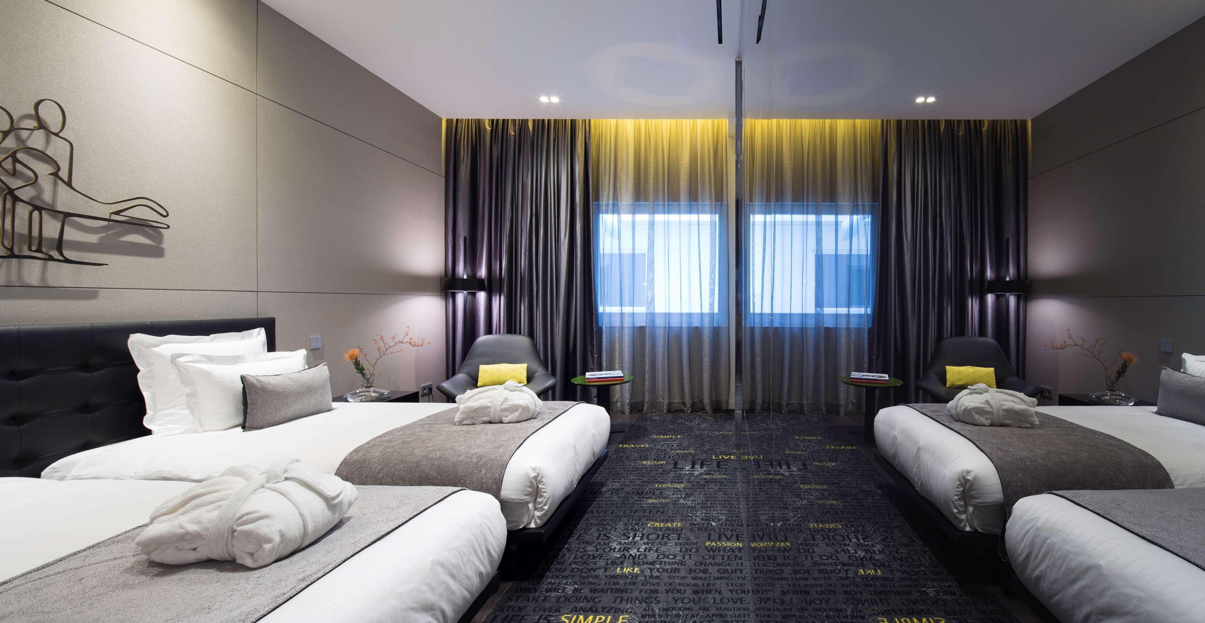 Une chambre d’hôtel moderne avec deux lits, une grande fenêtre avec des rideaux, une œuvre d’art accrochée au mur et une moquette noire ornée de citations inspirantes.