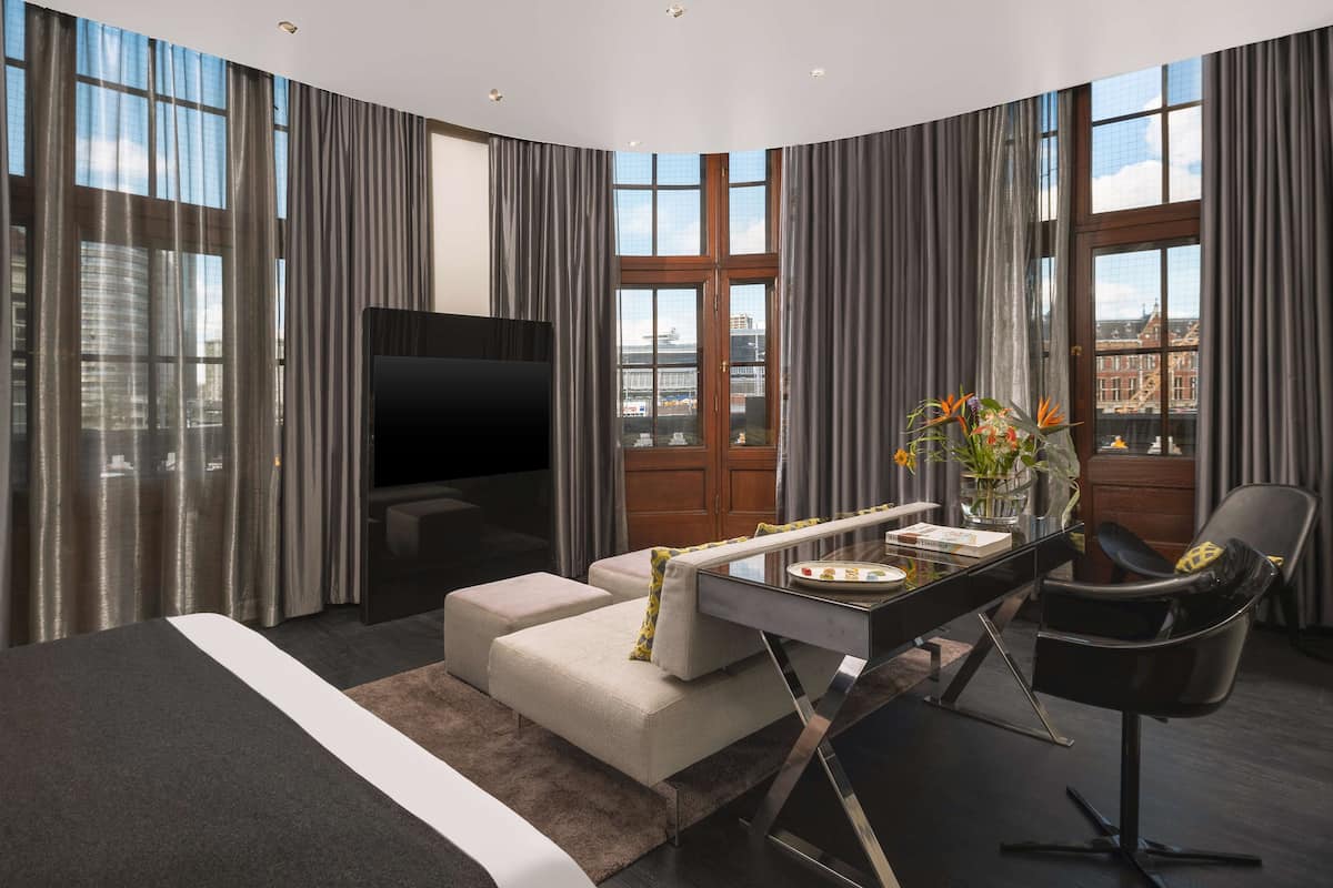 Ein modernes Hotelzimmer mit einem großen Bett, einer Sitzecke, einem Schreibtisch und einem Stuhl. Ein Fernseher ist an der Wand angebracht und es gibt ein Fenster mit Vorhängen.