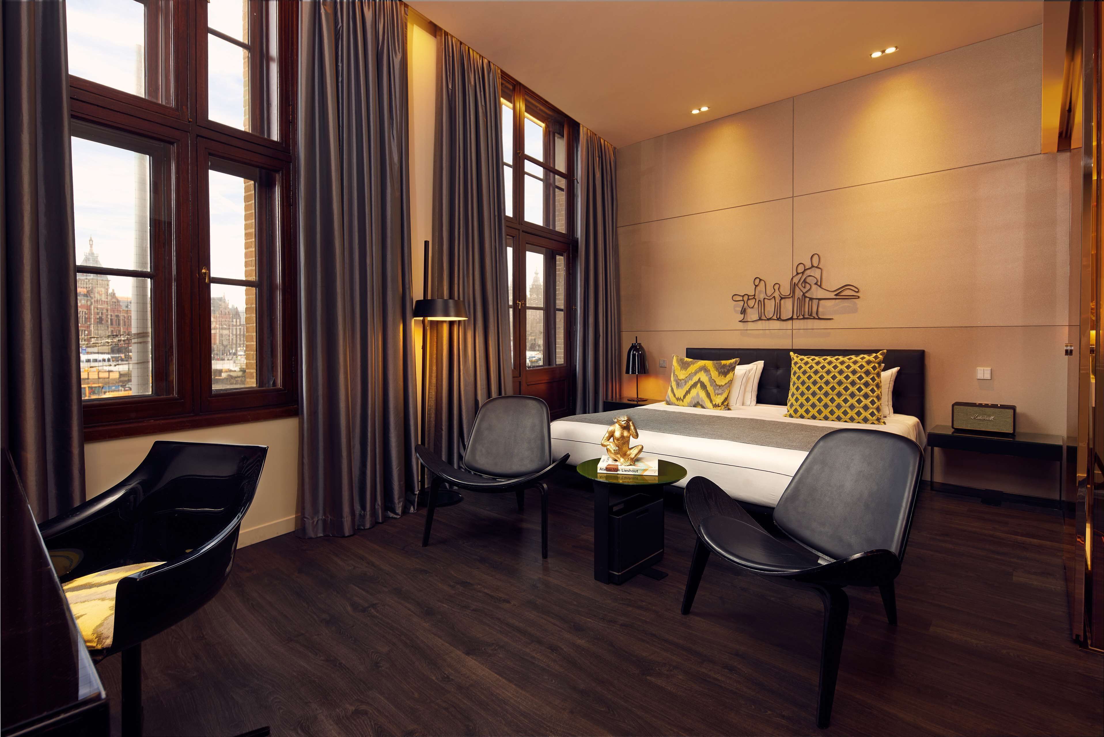 Habitación de hotel con una cama grande, dos sillones, una mesita y vistas a la ciudad a través de las ventanas.