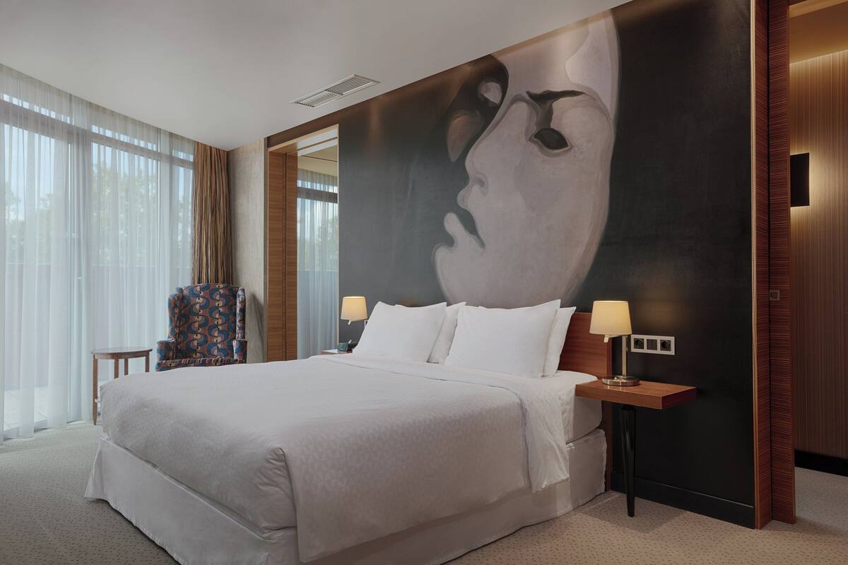 Une chambre d’hôtel moderne avec un grand lit, une table de chevet avec une lampe, une chaise et une grande fresque murale.