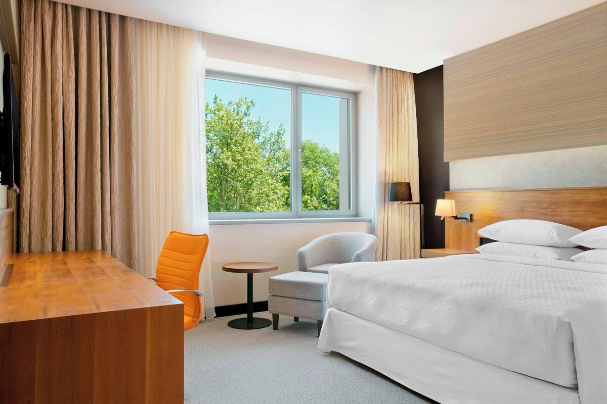 Une chambre d’hôtel avec un grand lit, un bureau, une chaise, une petite table et une fenêtre donnant sur des arbres.