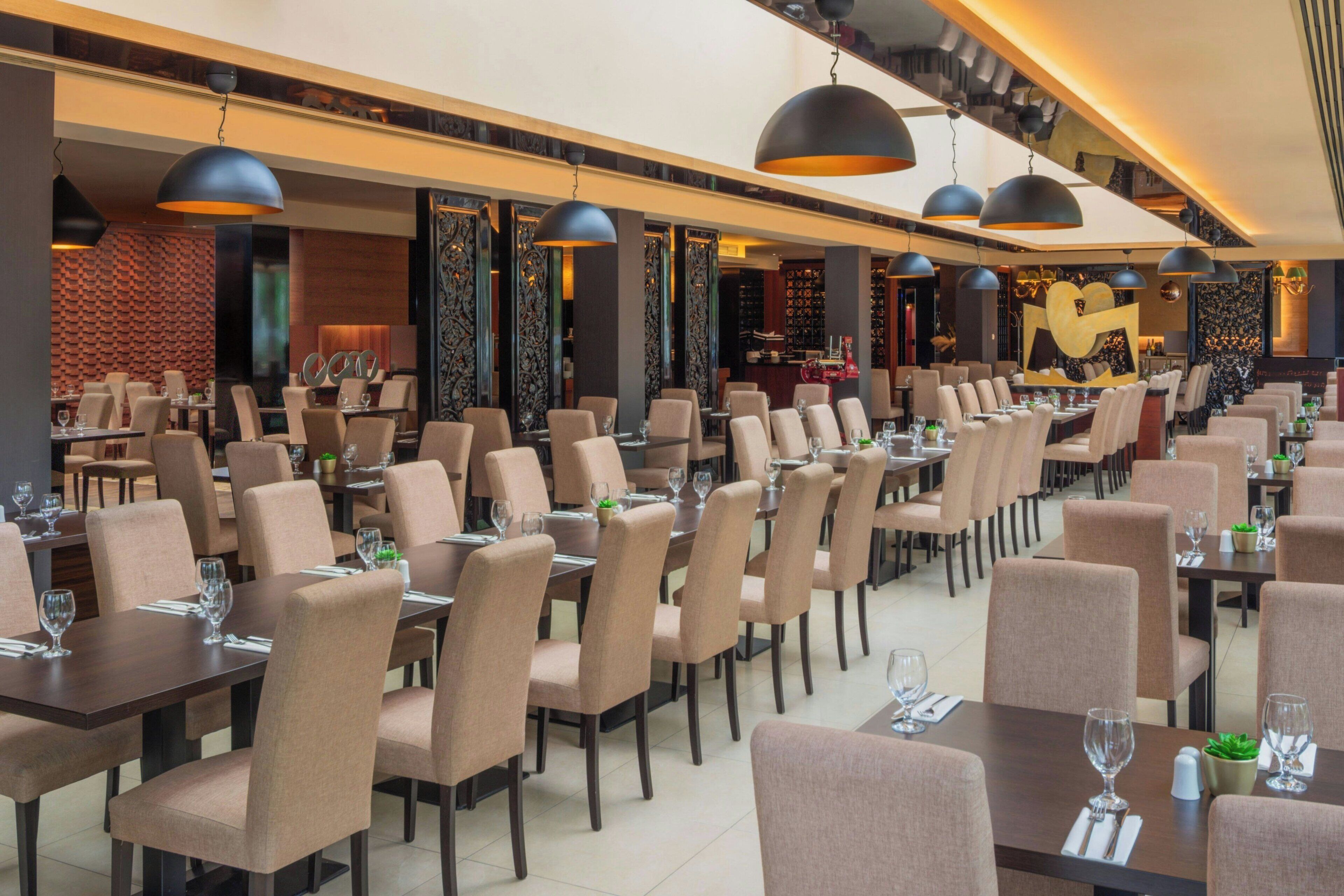 Un restaurante espacioso con numerosas mesas y sillas, grandes lámparas colgantes y una decoración interior moderna.