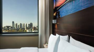 A hotel room with a bed, a nightstand, a TV mounted on the wall, and a window with a cityscape view.