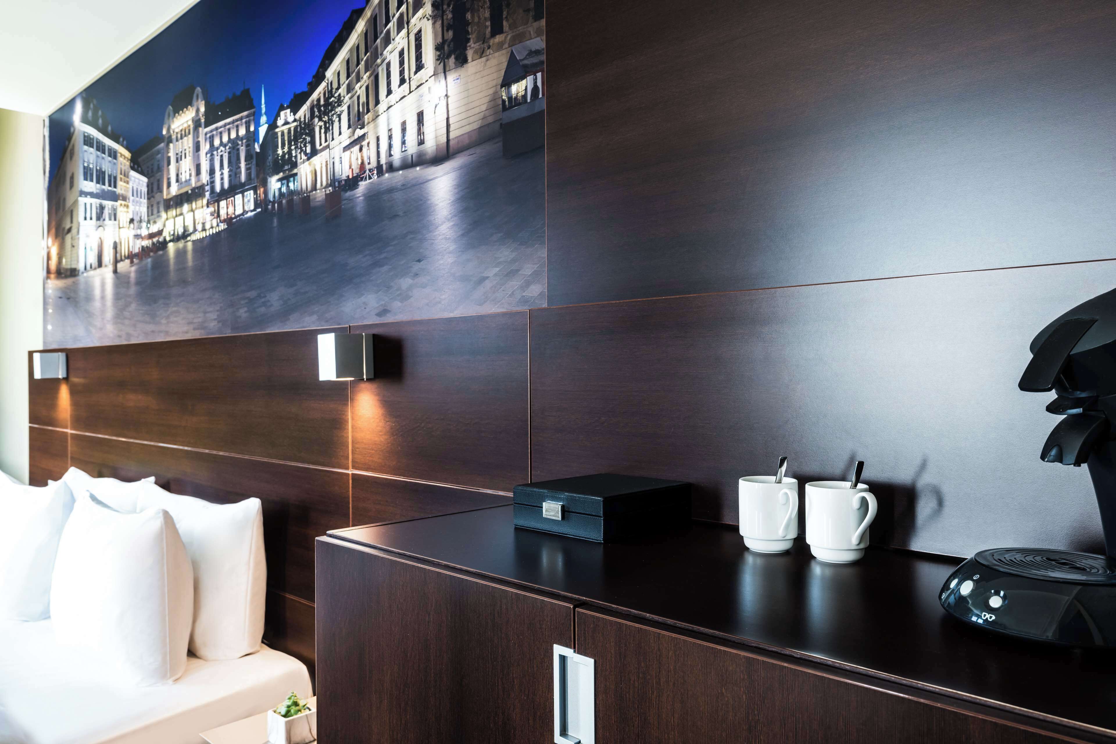 Ett modernt hotellrum med en stor målning av en stadsbild på väggen, en mörk träfärgad sänggavel, en vit soffa, en kaffemaskin och två vita koppar på bänkskivan.