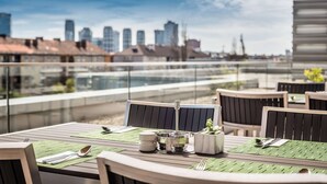 Un área de comedor en la azotea con mesas dispuestas para dos, vistas del horizonte urbano y una barandilla de cristal.