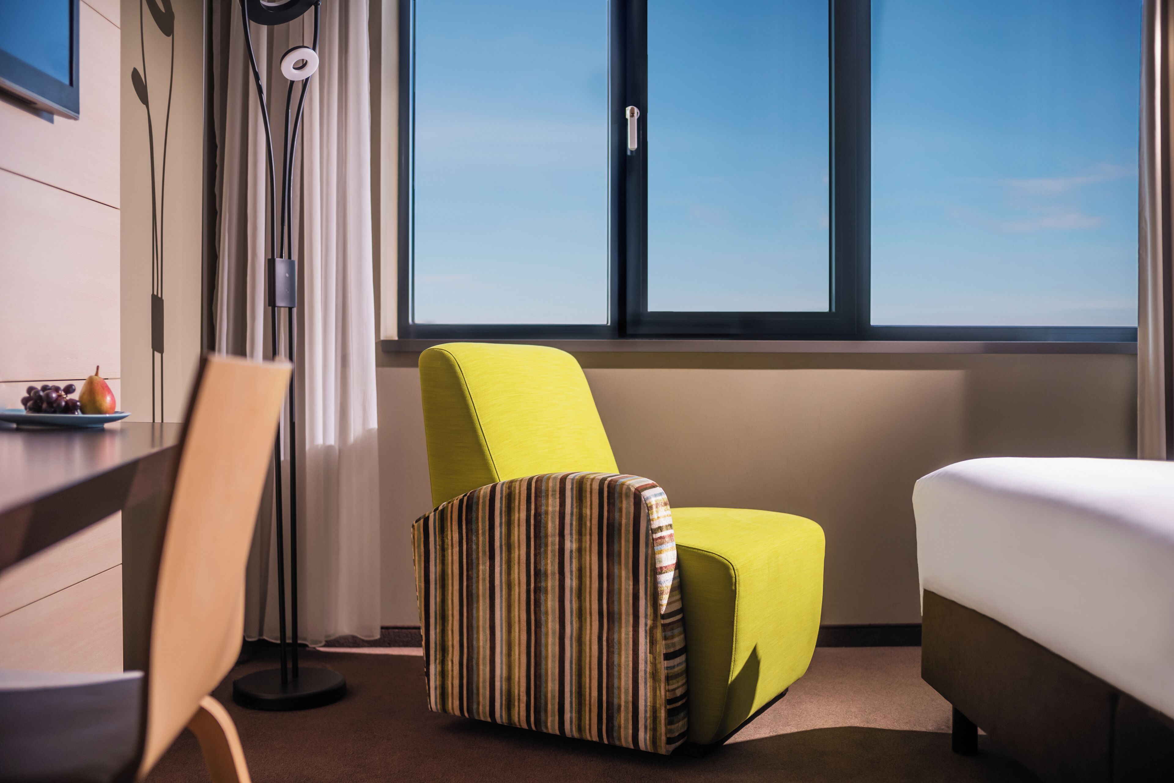 Ein Hotelzimmer mit einem Bett, einem grünen Sessel, einem gestreiften Hocker, einem Schreibtisch mit Obstplatte und einem großen Fenster mit Blick auf den Himmel.