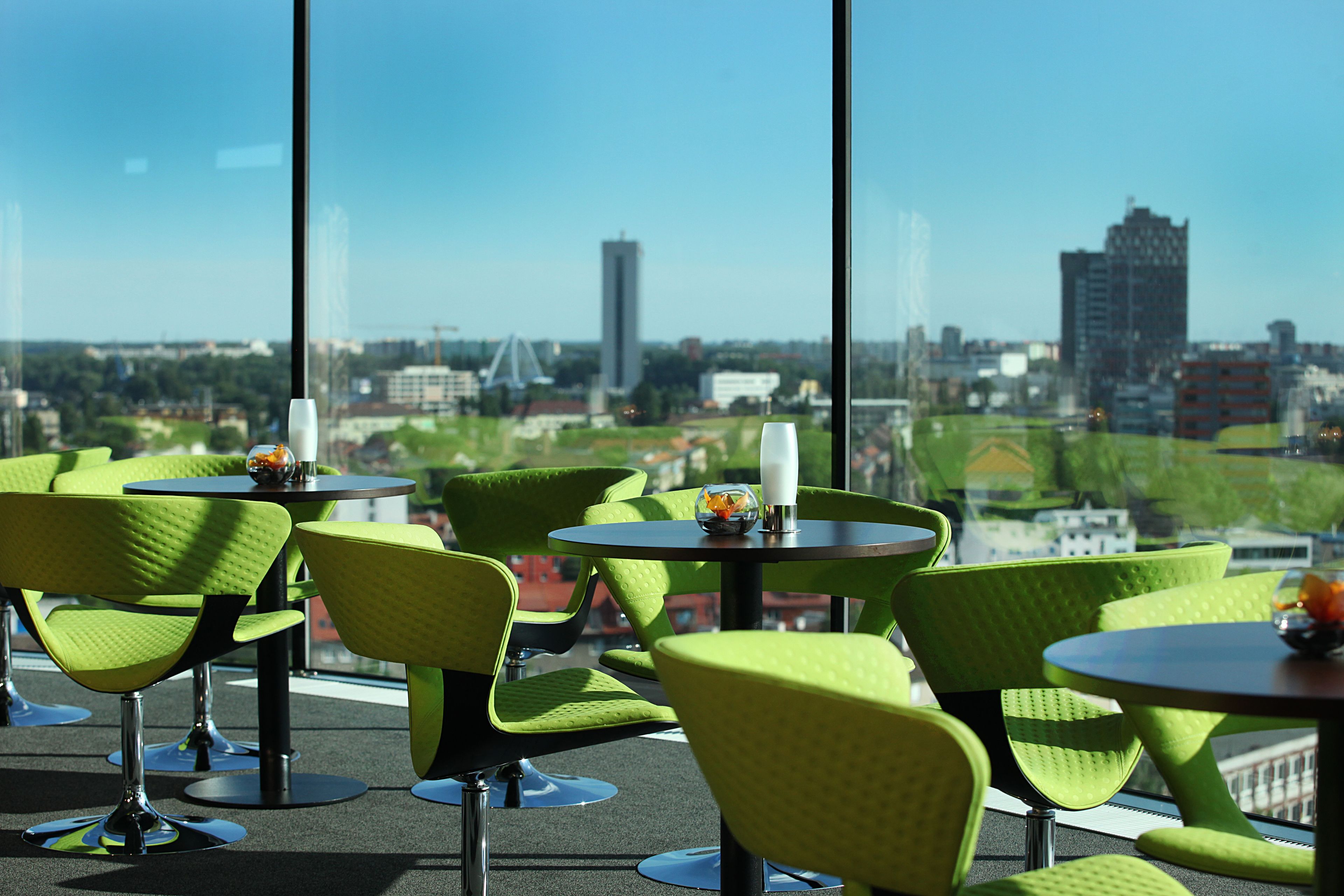 A modern indoor seating area with green chairs and tables, large windows offering a city view.