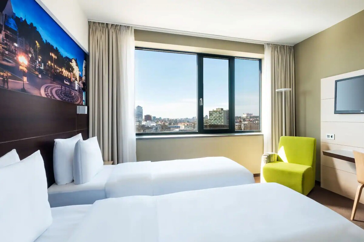 Habitació d'hotel amb dos llits, televisió, escriptori i finestra amb vista a la ciutat.