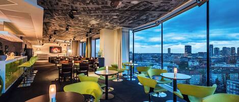 Un lobby de hotel moderno con vistas a la ciudad, asientos verdes y una zona de bar.