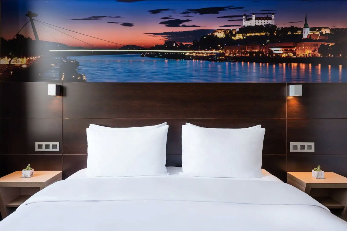 Habitació d'hotel amb un llit gran, dues taules de nit i un mural a la paret amb un paisatge urbà.
