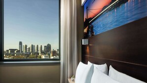 Habitación de hotel con un ventanal amplio que ofrece vistas a la ciudad, una cama bien hecha y un televisor de pared que muestra un atardecer sobre un puente.