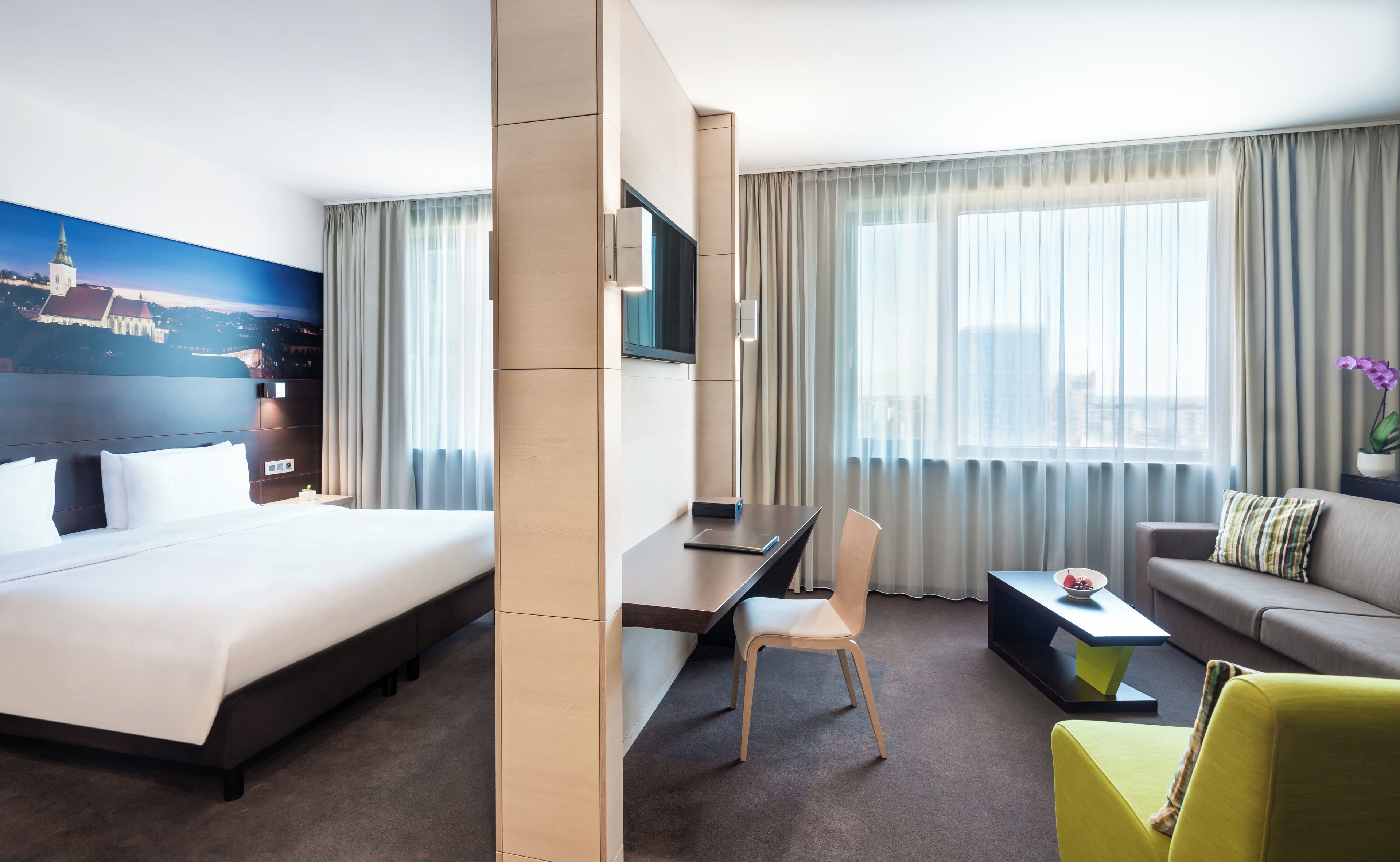 Habitación de hotel con una cama grande, un escritorio con una silla, un sofá, un televisor y un mural de paisaje urbano en la pared.