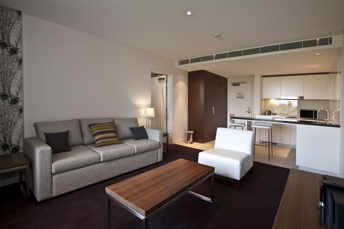 Un moderno salón con un sofá gris, una mesa de centro de madera y un sillón blanco. Se ve una cocina al fondo con armarios blancos y una encimera negra.