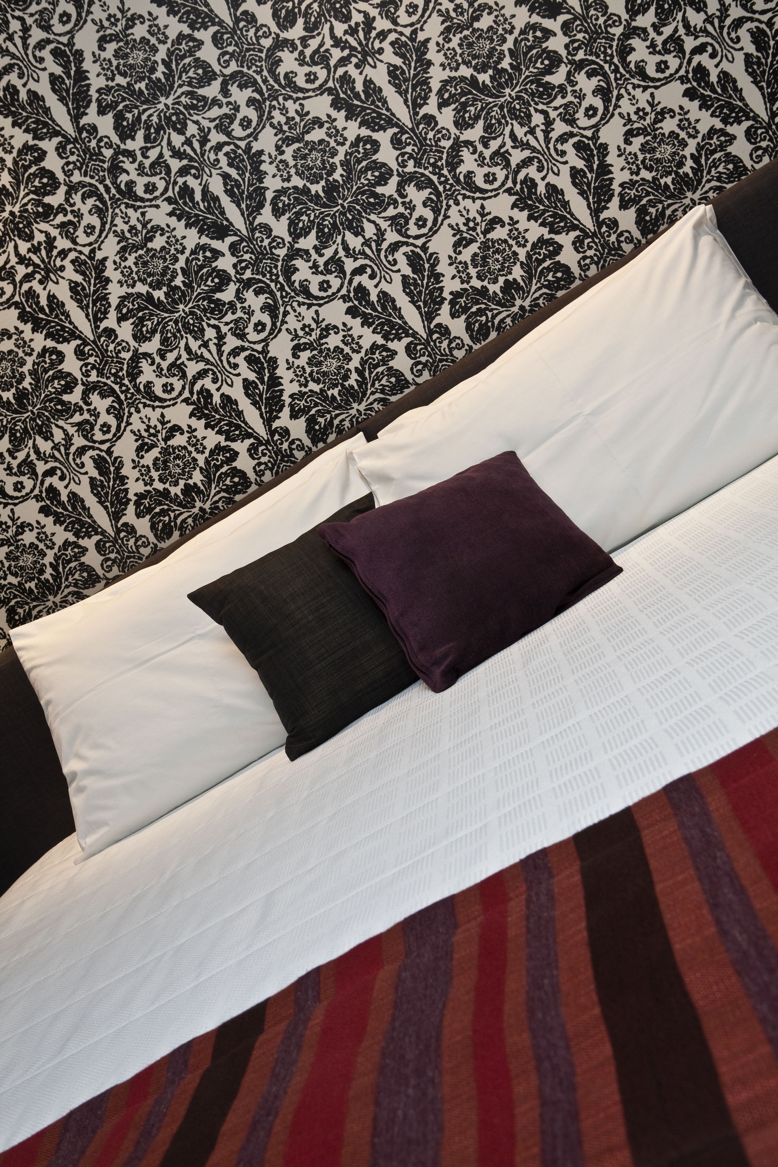 A bed with white bedding and two pillows, one black and one purple, against a patterned wallpaper.