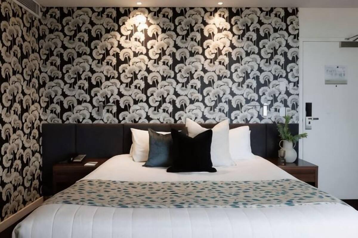 Un dormitorio con papel tapiz estampado, una cama con muchos cojines, una mesita de noche y una mesita auxiliar.
