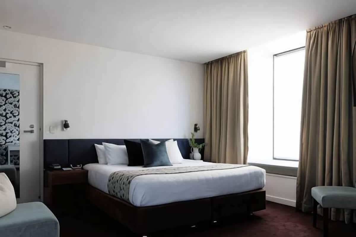 Habitación de hotel con cama, mesitas de noche, una silla, ventana con cortinas y puerta con diseño decorativo.