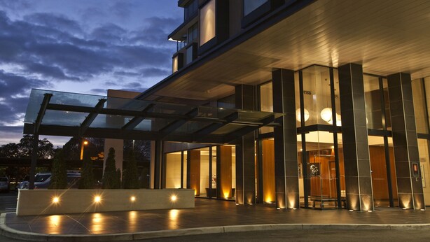 A modern hotel entrance with a glass canopy, a covered waiting area, and a well-lit lobby.