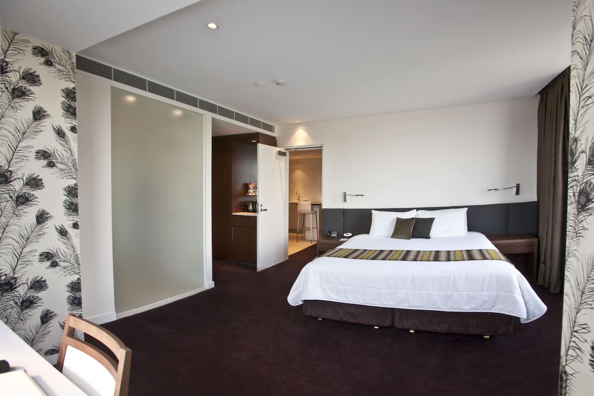Habitación de hotel con una cama grande, un escritorio y una silla. Hay una puerta que da a un baño y una kitchenette.