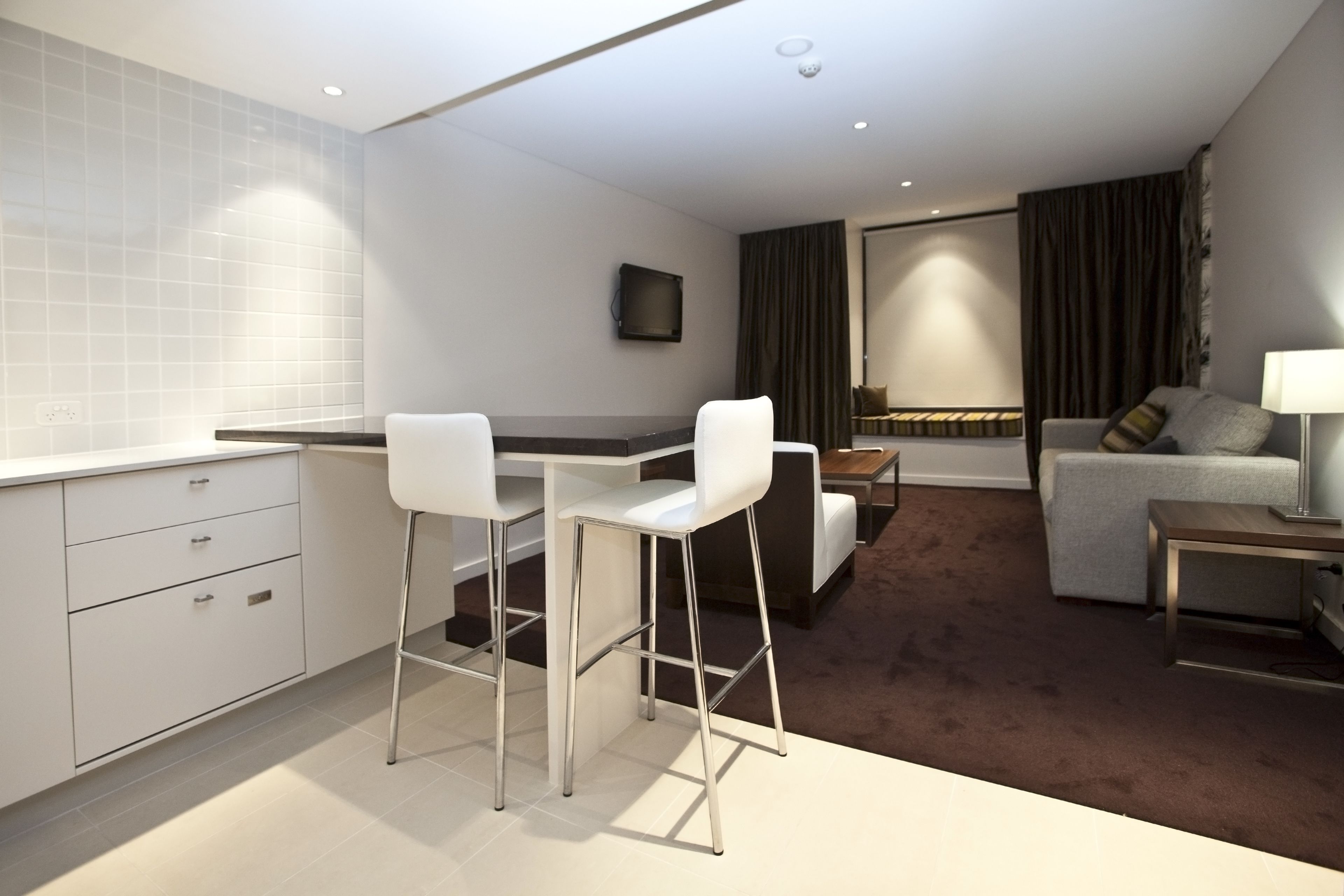 A modern hotel room with a kitchenette, a dining area with bar stools, a sofa, and a lamp.
