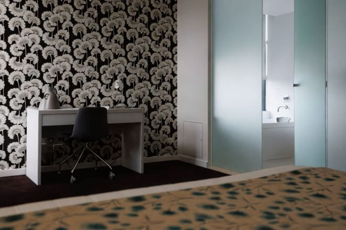 A room with a desk, chair, patterned wallpaper, a bed with a patterned bedspread, and a view of a bathroom.