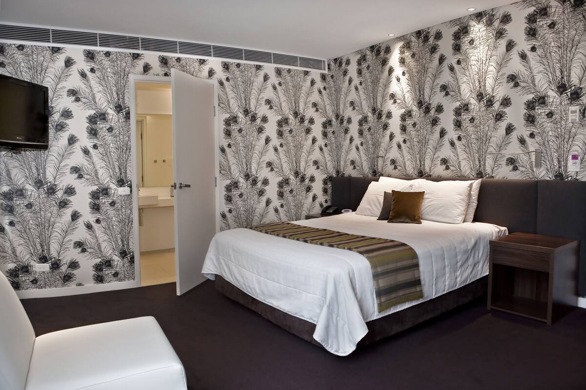Un dormitorio moderno con una cama grande, una mesita de noche, un televisor montado en la pared y una puerta que da al baño.