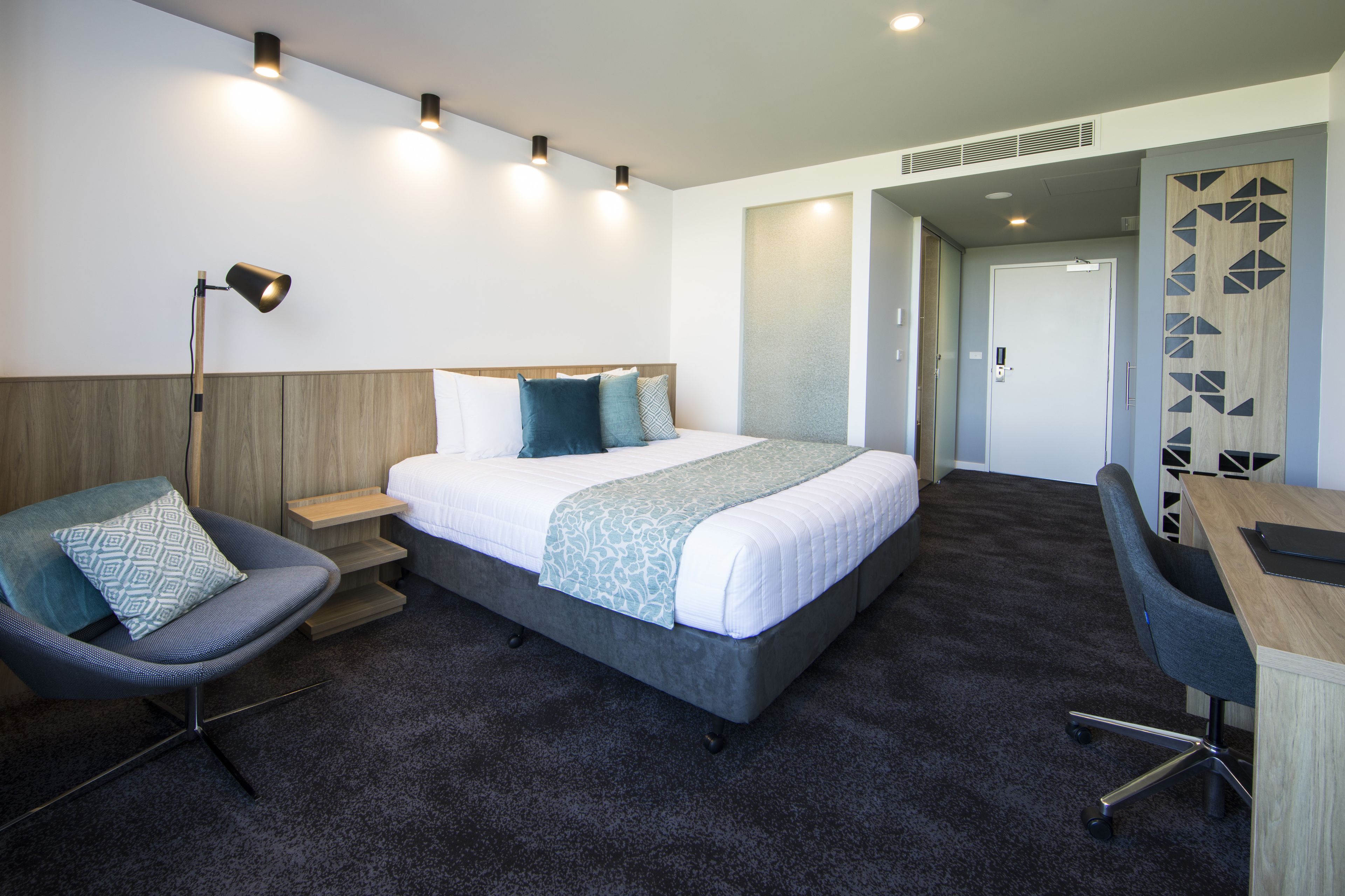 A modern hotel room with a large bed, a desk, a chair, and a lamp.