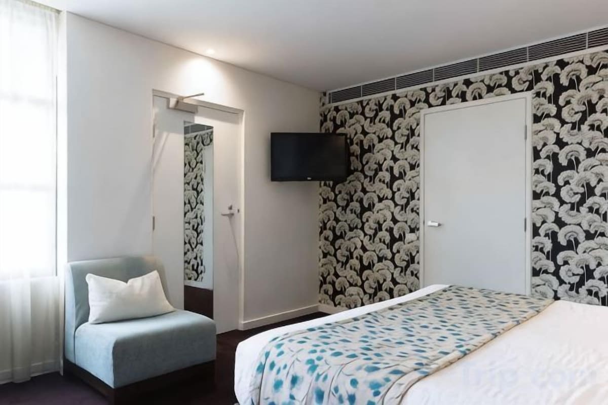 A hotel room with a bed, a chair, a television on the wall, and a door leading to another room.