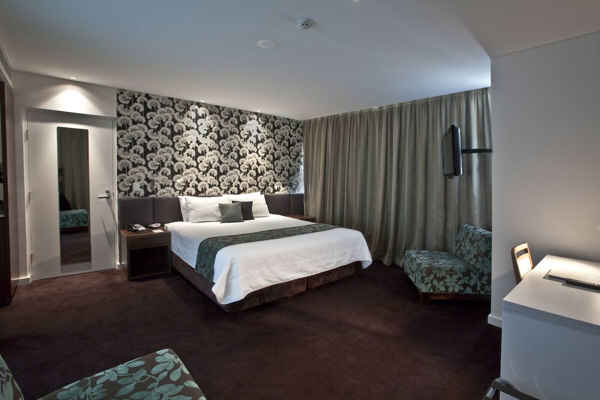 Habitación de hotel con una cama grande, una mesita de noche, un escritorio con lámpara, una silla y televisor montado en la pared.