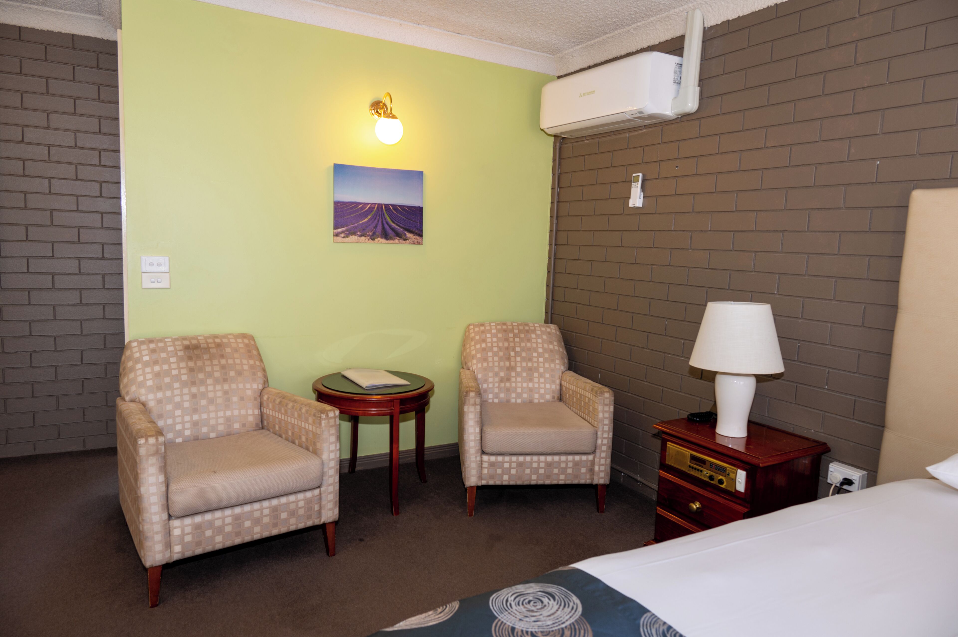 A hotel room with two armchairs, a small table, a lamp, a bed, and a wall-mounted air conditioner.