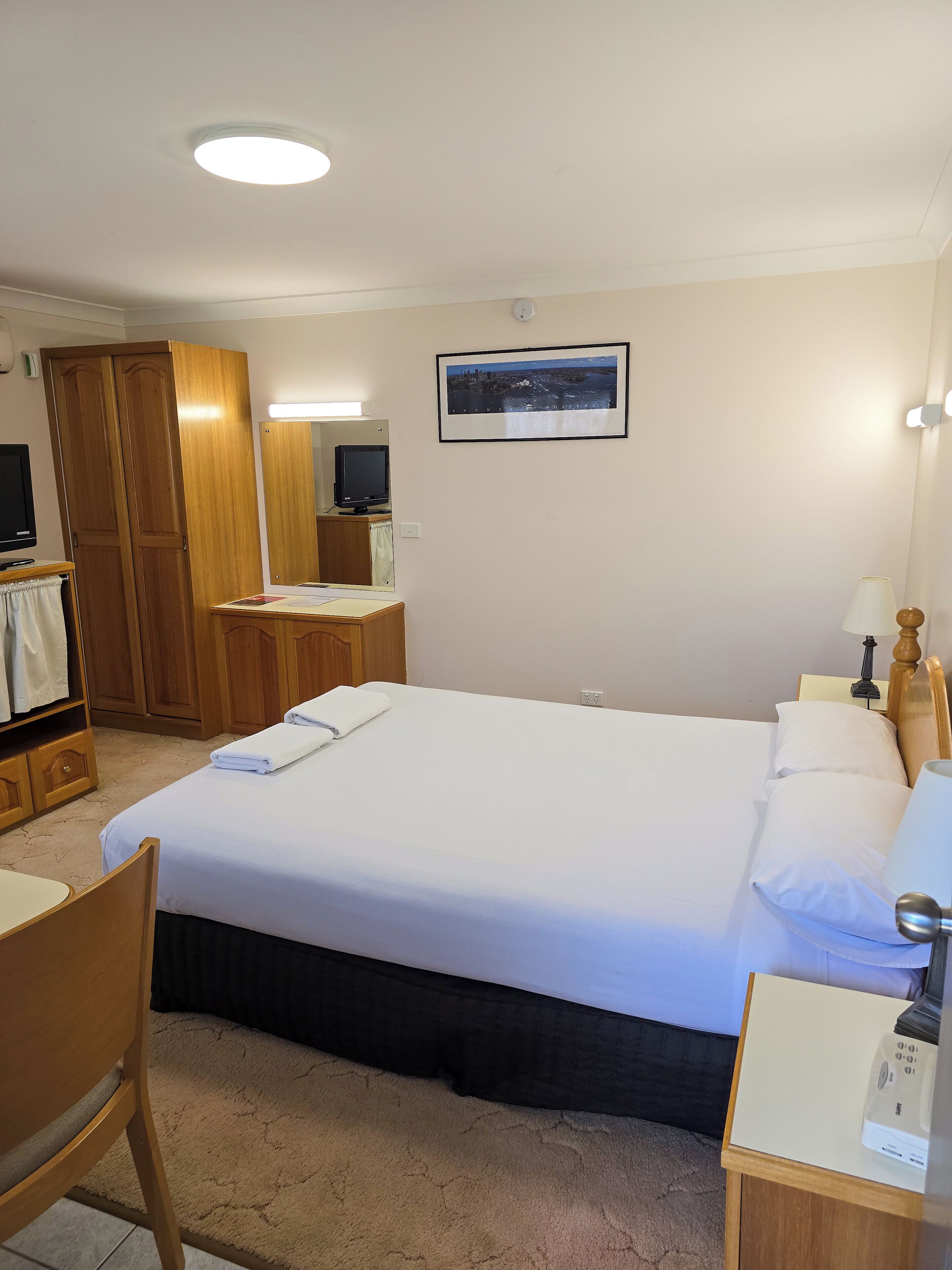 Quarto de hotel com uma cama grande, uma escrivaninha com computador, uma cadeira, um guarda-roupa e um quadro na parede.