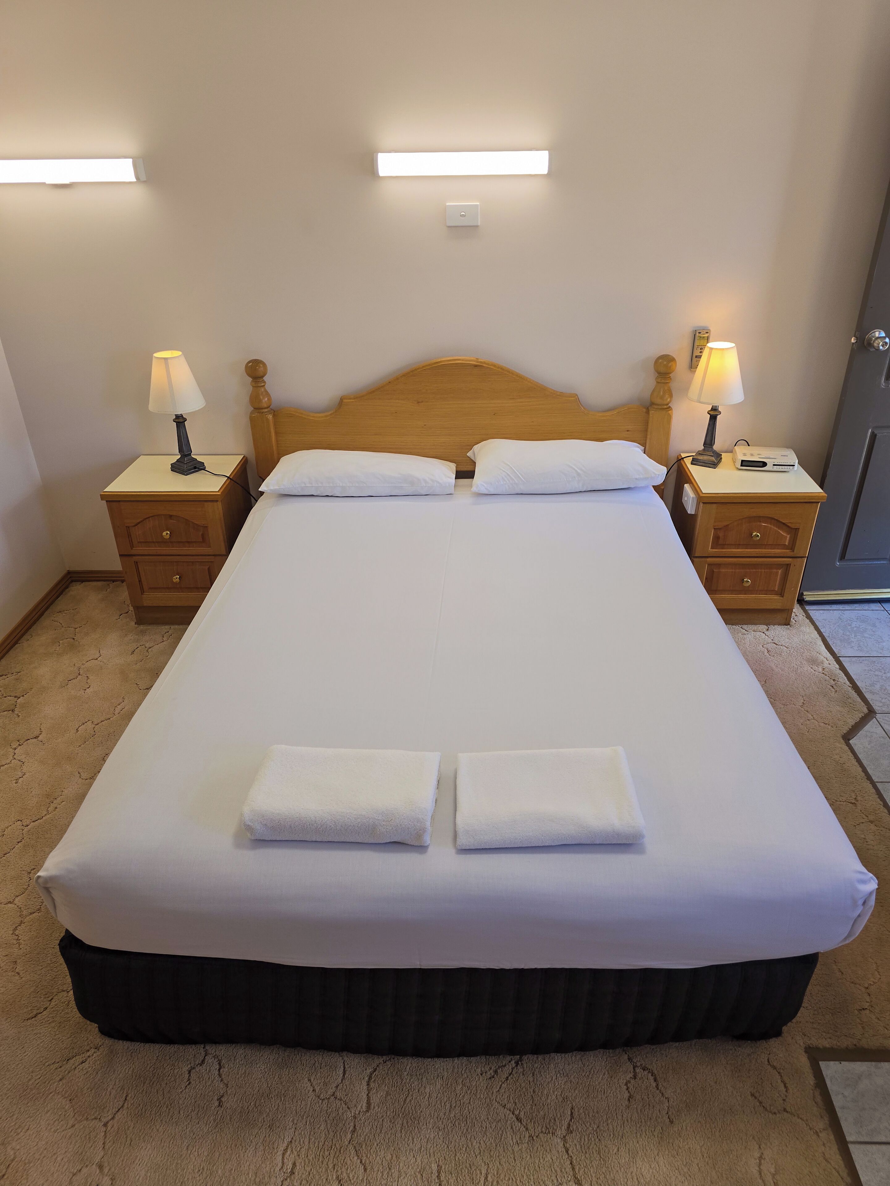 A neatly made bed with white linens and two folded towels, flanked by wooden nightstands with lamps, in a room with a wooden headboard and a plain wall.