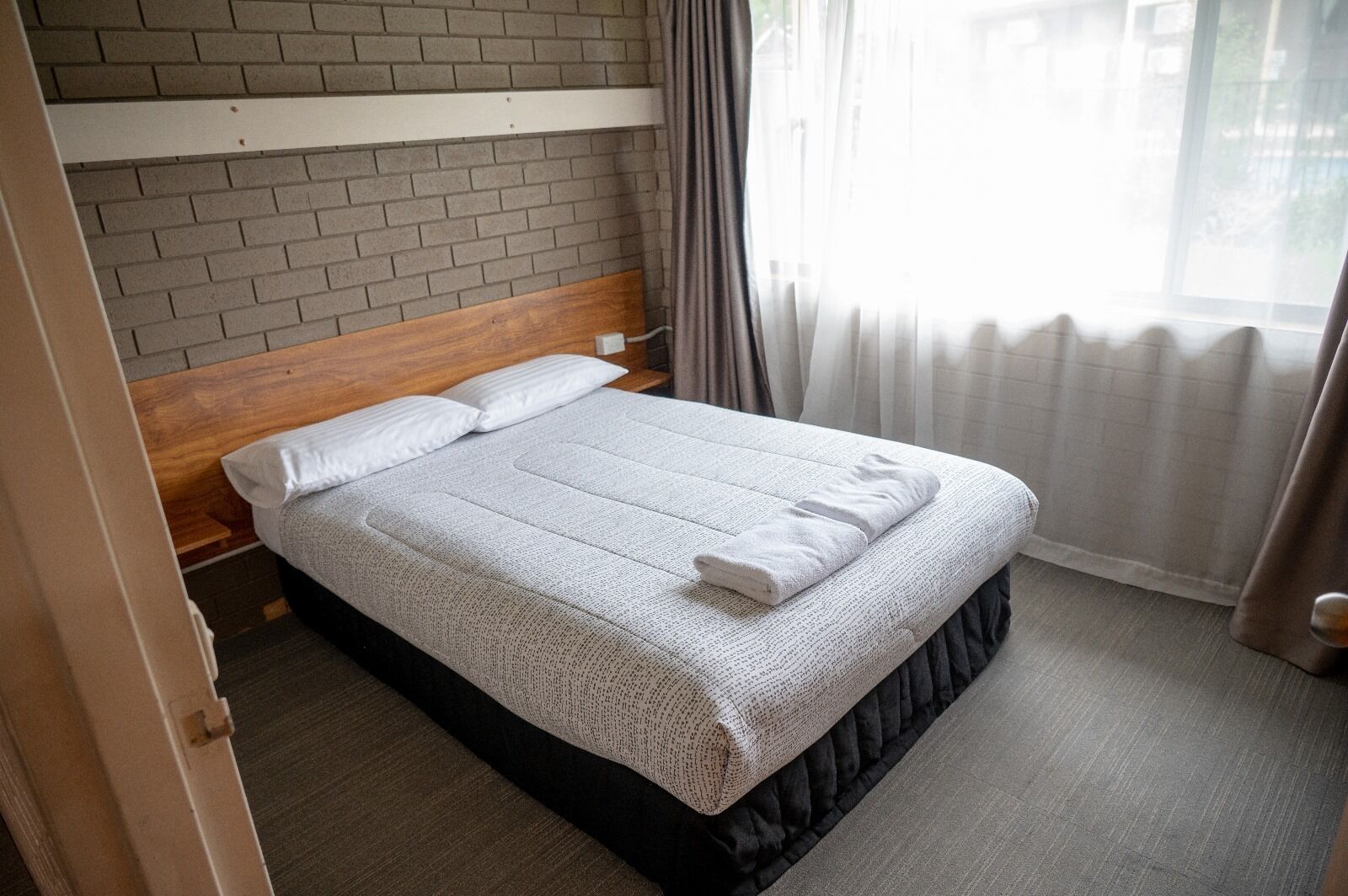 Un lit bien fait, avec une couette et des oreillers blancs, une tête de lit en bois et un mur en briques grises.