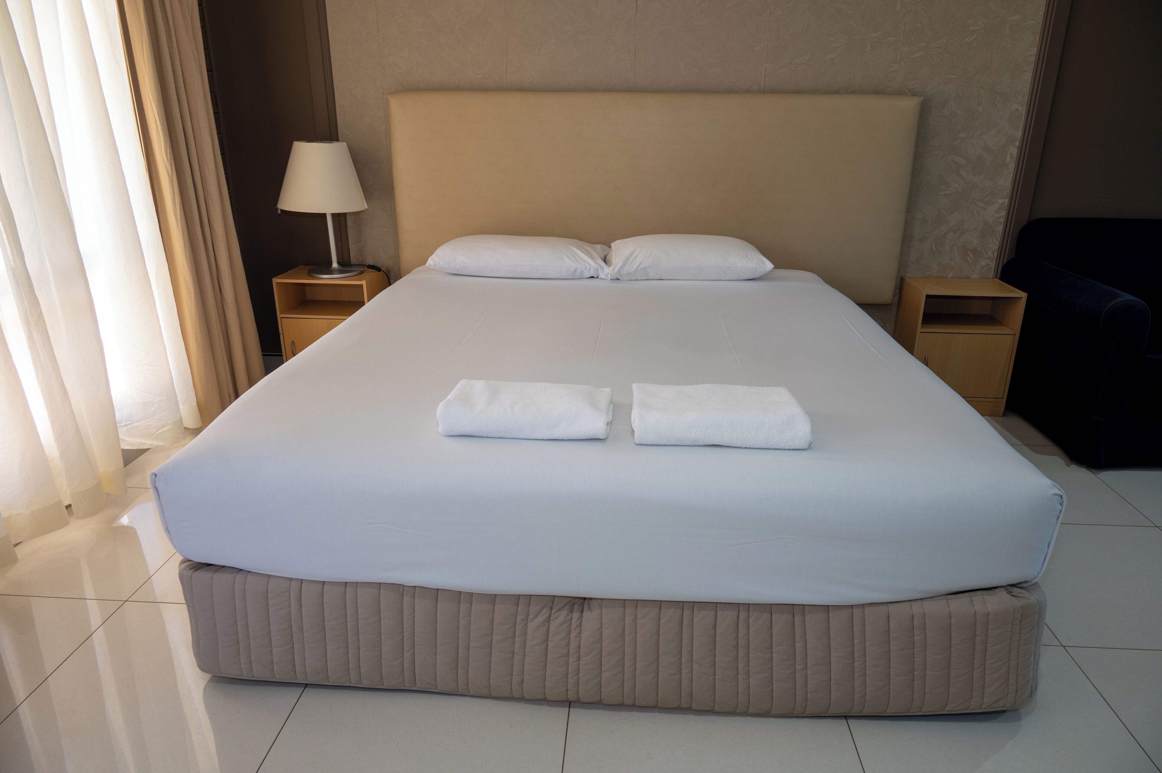 A neatly made bed with white linens and two folded towels, a bedside table with a lamp, and a dark-colored chair.