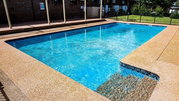 A rectangular swimming pool with a pebble-lined edge, surrounded by a brick building with air conditioning units.