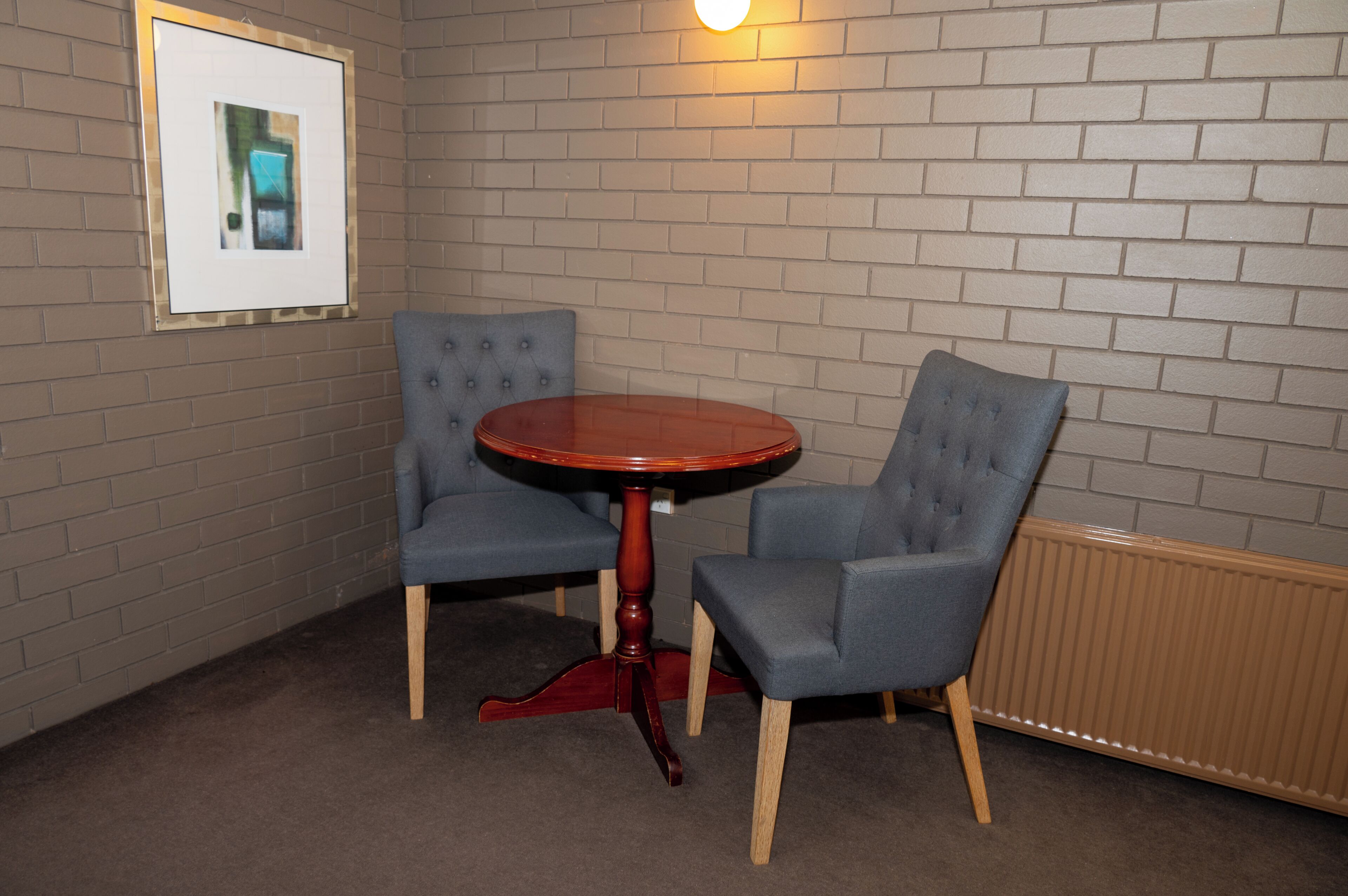 A room with a brick wall, a framed picture, a round wooden table, and two upholstered chairs.