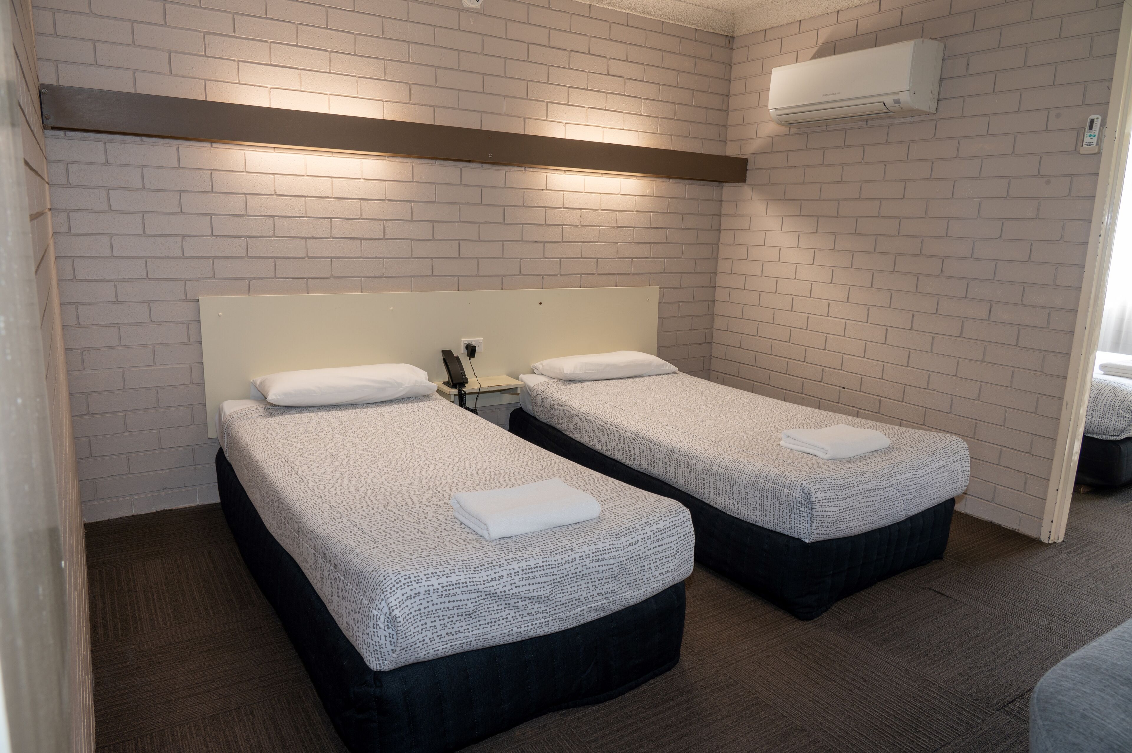 A hotel room with two beds, white brick walls, a ceiling-mounted air conditioner, and a wooden floor.